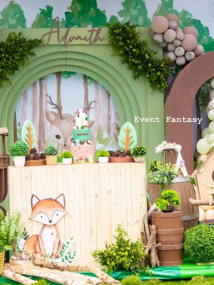 A beautiful woodland cake table for Advaith. The setup features a green arched backdrop with a forest illustration, a cute fox cutout, and various rustic elements like a wishing well and birdhouse.