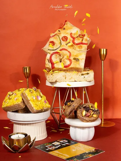 A creative shot of focaccia bread and other baked goods stacked on pedestals. The warm, orange background and falling petals create a dynamic and artistic image.