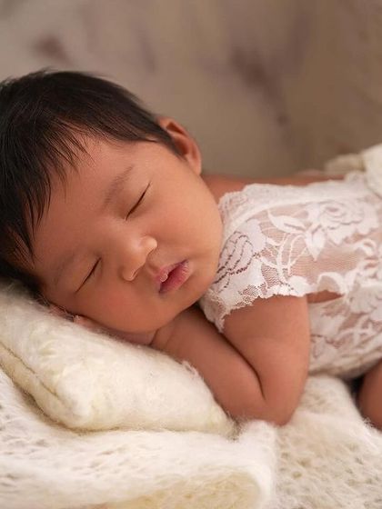 A close-up of a newborn in a white lace romper, sleeping peacefully on a soft pillow and framed by delicate white flowers.