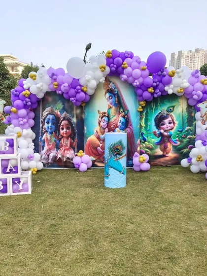 For this outdoor baby shower, we designed a beautiful and unique Krishna theme. The setup includes a large, vibrant backdrop with illustrations of Little Krishna, framed by a purple and white organic balloon arch, and custom 'BABY' blocks, all arranged perfectly on a lush green lawn.