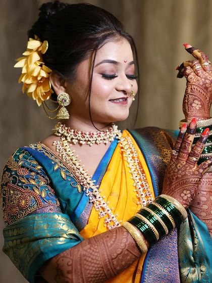 The bride's expression and pose highlight the beautiful henna design and the traditional green bangles. The makeup is kept soft and glowing to complement the vibrant yellow and blue saree.