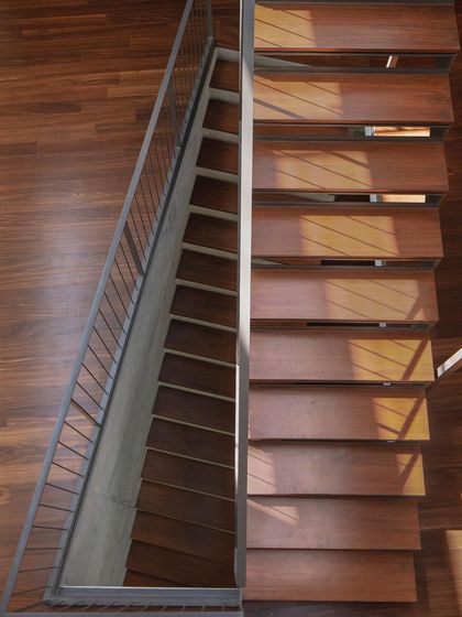 An aerial view of the staircase in the Debris Block House, highlighting the clean lines of the wooden treads and the play of sunlight filtering from above.