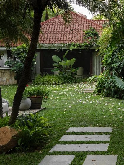 A tranquil backyard with a stone path leading across a manicured lawn towards a terracotta-roofed outbuilding. The garden is framed by palm trees and dense foliage, creating a feeling of a private, hidden escape.