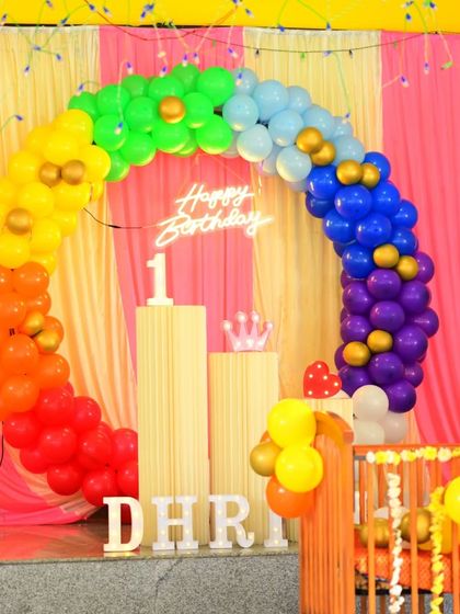 This vibrant rainbow-themed first birthday decoration features a full spectrum of colorful balloons in a circular arch. The setup is completed with a neon sign and pedestals for the cake.