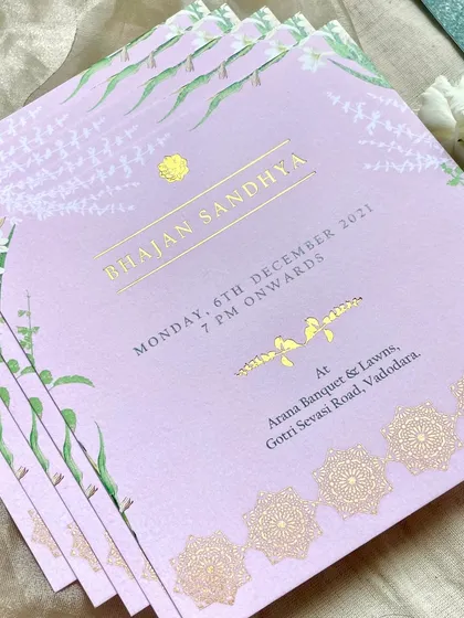 A stack of 'Bhajan Sandhya' invitations. The lilac color paired with gold foil and traditional mandala borders creates a design that is both serene and celebratory.