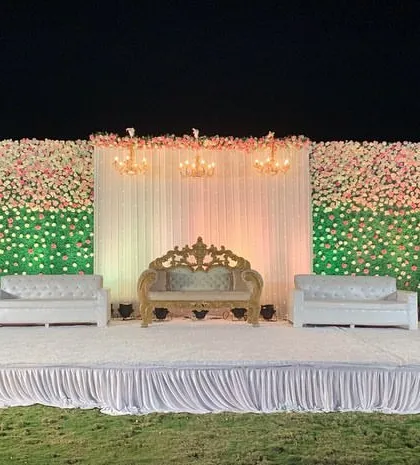 A wide, straight-on shot of a simple and elegant outdoor stage. The design features two large flower walls on either side of a central draped section with chandeliers.