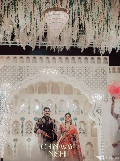 The couple's grand entrance against a stunning white backdrop with intricate jaali work and a floral ceiling. Cold pyro effects add to the magical moment.