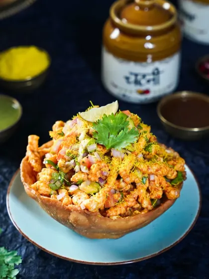 A crispy Tokri Chaat (basket chaat) filled with delicious toppings. The moody blue background and traditional pickle jars add a rustic yet refined feel to this popular street food dish.