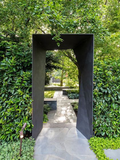 A modern, sculptural gateway provides a portal through a dense, living wall of ivy. This design contrasts the sharp, clean lines of the metal arch with the organic texture of the surrounding foliage.