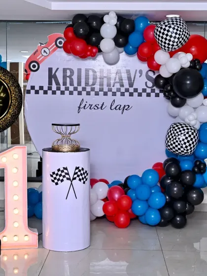"Kridhav's First Lap" race car themed birthday. This setup features a circular backdrop with checkered flag details, a red, blue, and black balloon garland, and a large light-up number '1'.