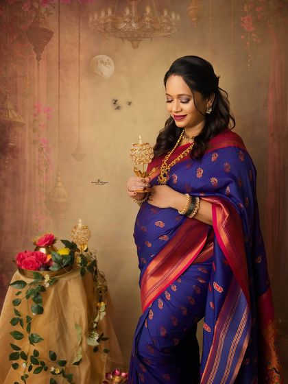 A solo maternity portrait capturing the festive spirit. The mother-to-be holds a traditional lamp, her beautiful blue saree glowing in the soft light of the Diwali-themed setup.