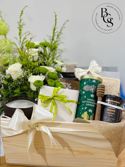 A nature-inspired gift hamper in a wooden crate with a burlap lining. This arrangement features fresh white and green flowers, a scented candle, cookies, and honey, creating a wholesome and calming gift.