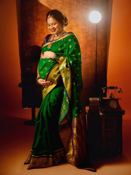 A full-length portrait showcasing the beautiful drape of a green silk saree. The soft lighting and vintage props add to the classic feel of the shot.
