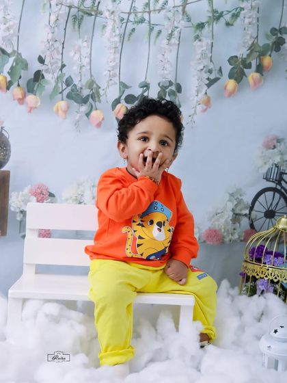 A cute toddler in an orange sweater sits on a white bench in our winter snow theme. The floral backdrop adds a touch of whimsical contrast to the snowy ground.