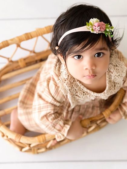 An overhead view of a baby girl with a floral headband looking up from a wicker basket, a classic sitter pose.
