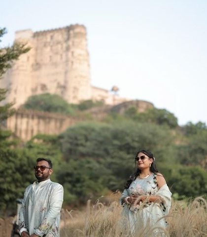 A wide shot capturing the couple in a field of tall grass with a historic fort in the background. This image combines natural beauty with the grandeur of Rajasthan's heritage.
