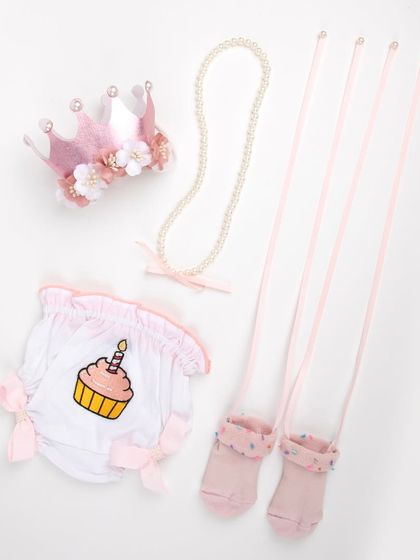 A baby's first birthday outfit set, including a cupcake-themed bottom, pink socks, a pearl necklace, and a floral crown.