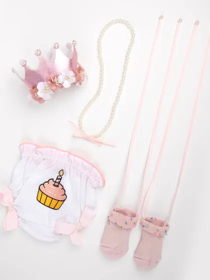 A baby's first birthday outfit set, including a cupcake-themed bottom, pink socks, a pearl necklace, and a floral crown.