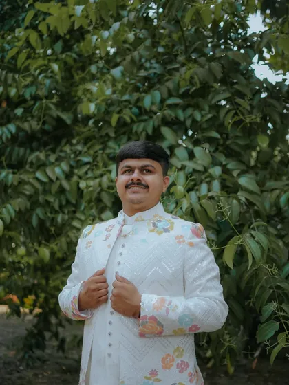 A classic portrait of the groom, looking dapper in his floral-embroidered bandhgala jacket against a natural green background.