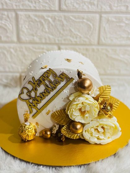 An elegant anniversary cake in white and gold. This design is adorned with golden flowers, leaves, and a Ferrero Rocher for a touch of sweetness.