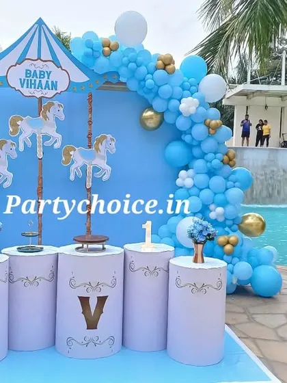 A charming carousel-themed party by the poolside. This setup features a custom blue backdrop with carousel horse cutouts, a beautiful blue, white, and gold balloon arch, and themed pedestals for desserts and cake.