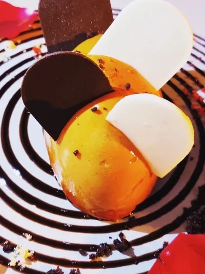 For sit-down dinners, I create artistic plated desserts like this Mango Chocolate Ecstasy. It features a mango mousse sphere adorned with dark and white chocolate, served on a plate with a mesmerizing chocolate swirl design.