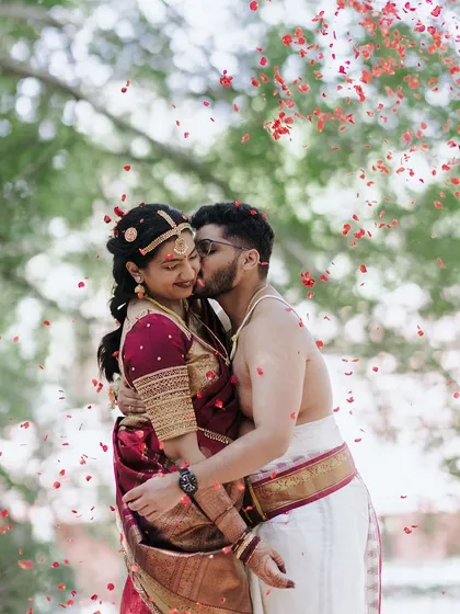 A tender kiss amidst a shower of red petals. The natural greenery provides a stunning contrast, making this a truly passionate and memorable wedding photograph.
