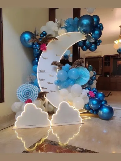 A dreamy moon-themed backdrop for a party. The crescent moon prop is lit from behind and surrounded by a cloud of blue and white chrome balloons.