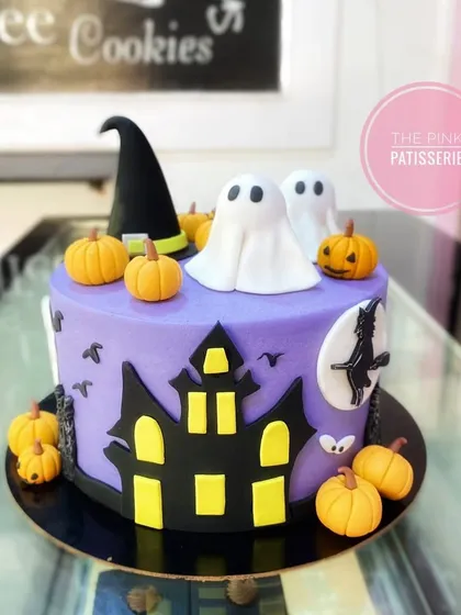 A spooky and fun Halloween cake. This purple cake is decorated with a haunted house silhouette, fondant ghosts, pumpkins, and a witch's hat.