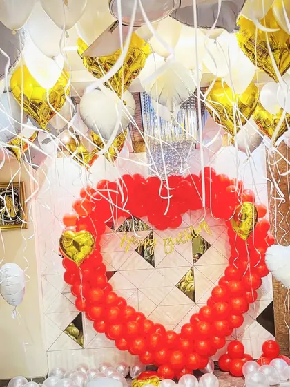 A grand romantic gesture featuring a large heart-shaped frame made of red balloons. The room is filled with white and gold balloons, perfect for an anniversary or proposal.