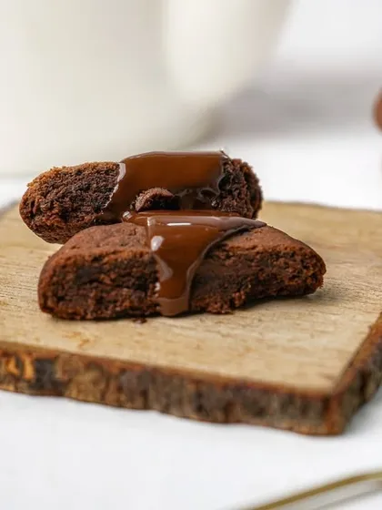 A soft chocolate cookie broken in half with chocolate sauce drizzled on top, styled on a rustic wooden board. This shot emphasizes the gooey, decadent texture of the cookie.