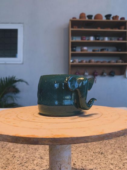 Another view of the deep green elephant mug, showing off its glossy glaze and charming details. In the background, you can see shelves of other creations waiting in my studio.