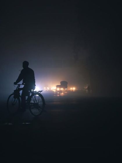 A cyclist is silhouetted against the headlights of distant traffic on a foggy night, a lonely but beautiful urban scene.