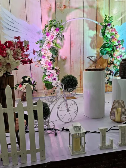 A whimsical and angelic-themed decoration. This creative setup includes floral arches adorned with large white angel wings, a decorative white bicycle, and a picket fence for a fairytale feel.