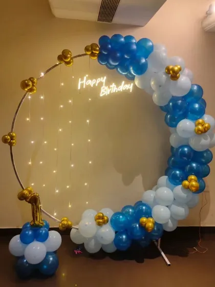 A simple and sweet setup for a first birthday celebration. This design uses a metal ring frame decorated with blue, white, and gold balloons, a small 'Happy Birthday' neon sign, and a number '1' foil balloon.