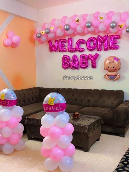 A simple yet charming welcome home setup. The decor includes a pink and white balloon garland with "Welcome Baby" foil letters and two "It's a Girl" balloon stands, perfect for a cozy celebration.