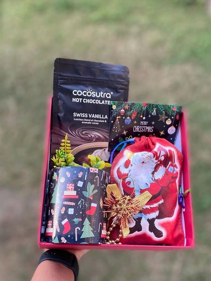 A festive collection designed to spread holiday cheer. This hamper includes a Christmas mug, hot chocolate mix, and a Santa-themed pouch filled with surprises, all held in a bright red gift box.