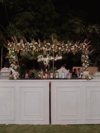 A chic and stylish bar setup for an outdoor Sangeet, featuring a 'Drunk in Love' neon sign and a floral arch with pampas grass.