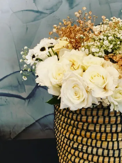 A close-up of a rustic-chic floral arrangement. The combination of white roses and dried baby's breath in a woven basket is perfect for adding a touch of elegance to any home occasion.