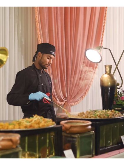 A chef at a live station preparing a noodle dish amidst an elegant setup with decorative vases and soft lighting.