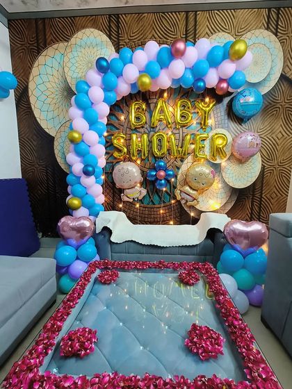 A lovely baby shower setup in a home setting. The decor features a blue and pink balloon arch over the sofa, with rose petals on the table, creating a cozy yet celebratory atmosphere.