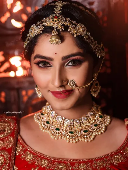 A classic bridal close-up. The focus is entirely on the bride's face, her expressive eyes, and the stunning details of her maang tikka and necklace. The warm, fiery background adds to the intensity.