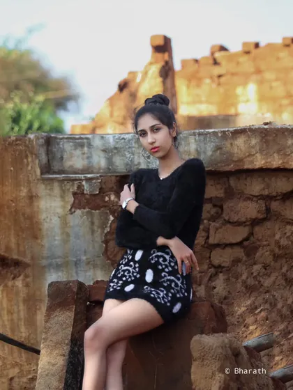 A high-fashion shot in a rustic, outdoor setting. The model wears a black sweater and a patterned mini-skirt, creating a strong contrast with the earthy, dilapidated background.