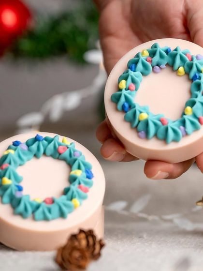 A lifestyle shot of a hand holding a Christmas wreath-shaped soap. This helps show the product's scale and connects it to the idea of holiday gifting.