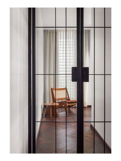 A glimpse through a metal pivot door reveals a lounge area with a custom-designed caned armchair. I use furniture to create inviting destinations within a home, encouraging moments of quiet relaxation.