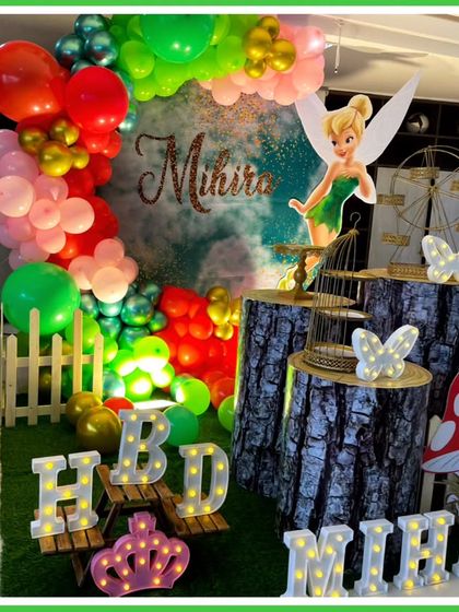 A whimsical fairy-themed third birthday party. The decor includes a Tinkerbell cutout, a forest backdrop, log-style plinths, and a large light-up number '3', creating an enchanted woodland scene.