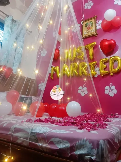 A romantic 'Just Married' bedroom decoration with a fairy light canopy, rose petals, and red and white balloons.