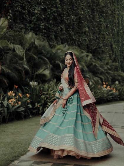 The bride in her vibrant Sangeet lehenga, a beautiful blend of turquoise, peach, and red. This portrait, set against a lush green background, showcases her radiant and colorful personality.