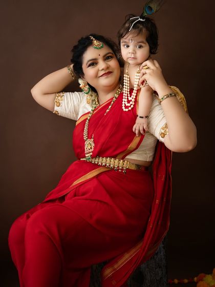 A beautiful portrait of another mother and child celebrating Janmashtami. The mother's red saree and the baby's pearl jewelry stand out against the simple brown backdrop.