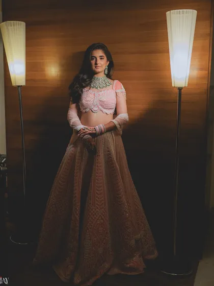 A striking solo portrait of the bride. She stands between two modern floor lamps that frame her perfectly, highlighting her beautiful pink lehenga and confident pose.